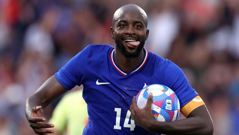Mateta, in France's blue kit and with his tongue sticking out of his mouth, celebrates with the ball under his left arm after scoring against Spain in the 2024 Olympics men's football final