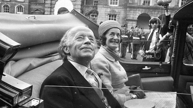 Erskine and Rita sitting in a car smiling. The roof is down in the vehicle and they are looking up. Erskine has a suit on and has short hair. Rita has shoulder length hair and is wearing a hat with a scarf, coat and gloves.