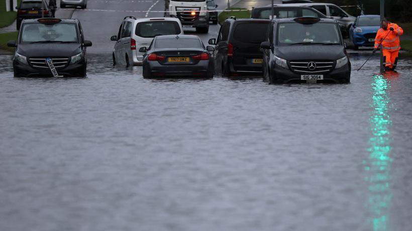 A council worker attempts to unblock drains after heavy overnight rain caused roads to flood, leaving cars stranded in Manchester