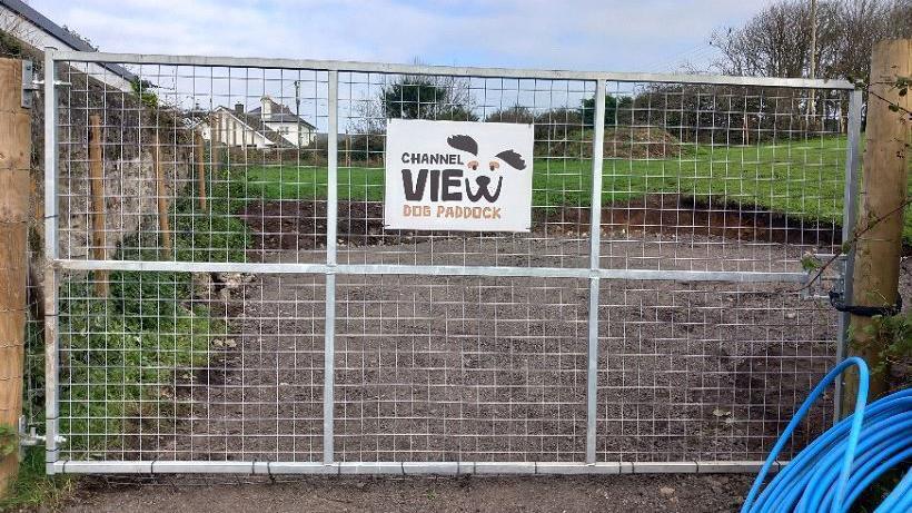 The picture shows a chain link fence to a grass field with the company's logo on a white sign in the middle. There are wooden posts either side and what appears to be some blue water piping on the floor. There are some fields and some houses in the background. 