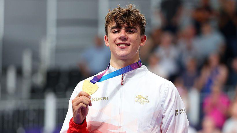 A smiling Henry Hobbs wearing a red and white Team England trackshuit shows his gold medal to the camera