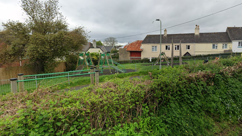 A picture of the play park in Kingsteignton. It is a large park with an area for children to play. It is surrounded by metal fences and hedges around the area.