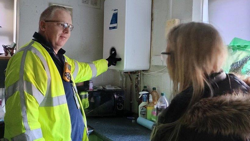 A gas engineer wearing a high-visibility jacket and goggles in a kitchen with a woman facing away from the camera. 