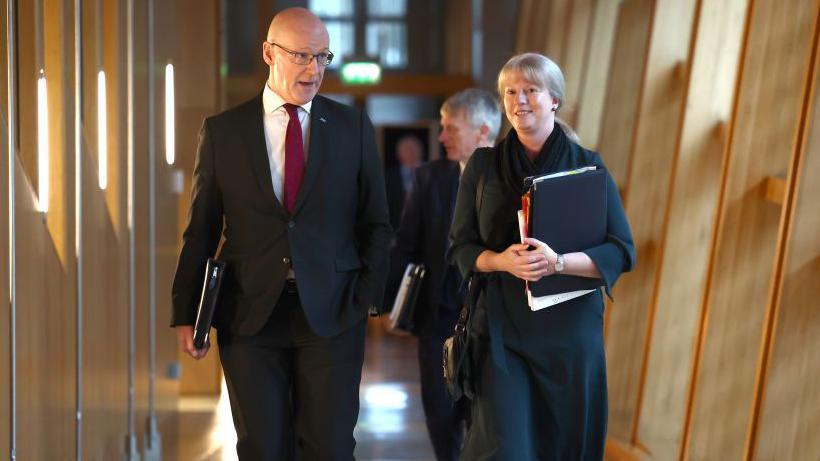 John Swinney, a bald man with glasses wearing a suit and burgundy tie walking down a corridor in Holyrood. He is carrying a folder and is joined by Shona Robison. She has blonde hair tied back in a ponytail and is also carrying a folder.