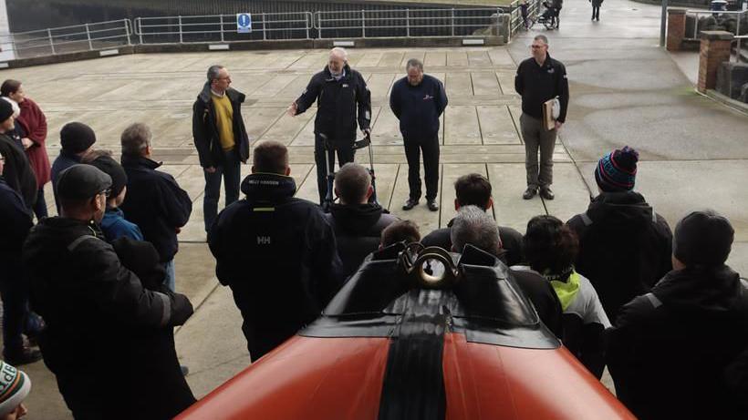 Two men wearing navy blue fleece jackets are seen standing outside of a lifeboat station in Portishead accepting an award. A group of people are standing in front of an orange lifeboat that is pictured.