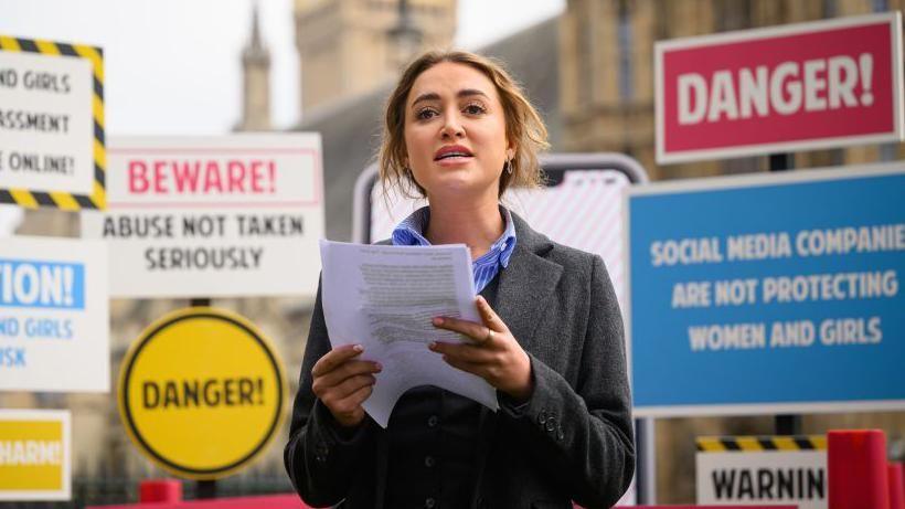 Georgia Harrison, a blonde woman wearing a blue and white stripy shirt and a grey coat, holding a piece of paper and making a speech in front of a selection of signs warning that social media companies do not take reports of abuse against women and girls seriously.