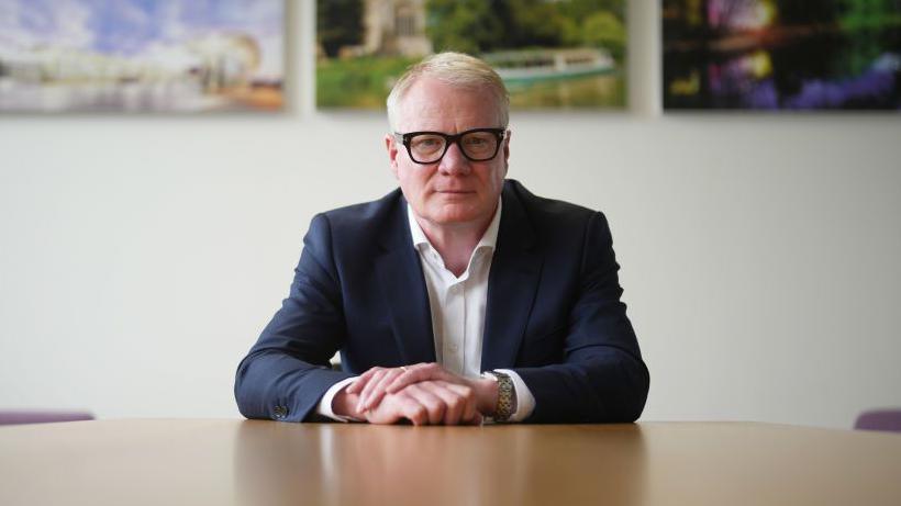 A man with pale blonde and white hair and dark rimmed glasses sitting alone at a table, looking straight at the camera. He is wearing a dark suit jacket and white shirt and has a serious expression on his face. A white wall with pictures of landscape scenes can be seen out of focus behind him.