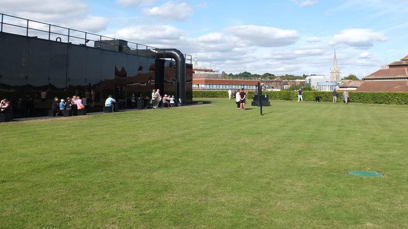 Staff members on a roof garden. There is a very large green lawn and the view is of the tops of other buildings including a church spire. 