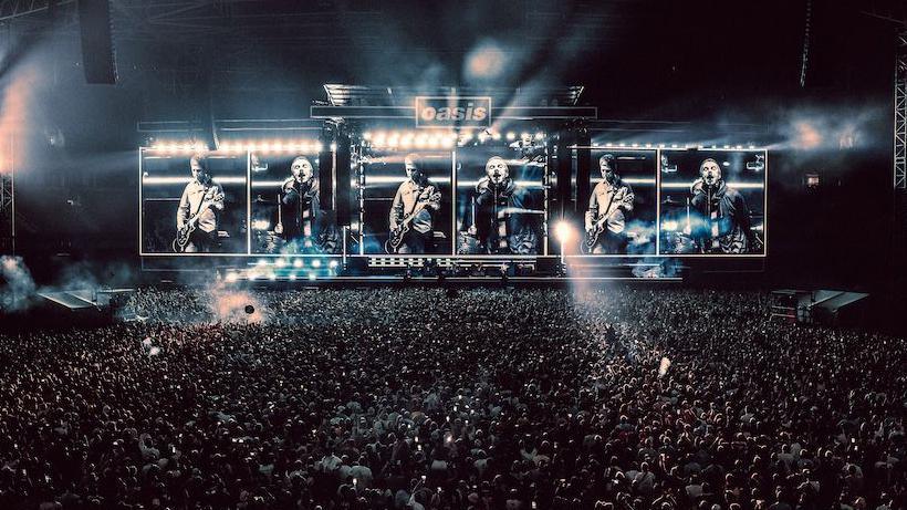 Wide shot of Oasis on stage in Cardiff. Thousands of people can be seen standing in front of the stage. Pictures of Liam and Noel performing can be seen on the stage screens.