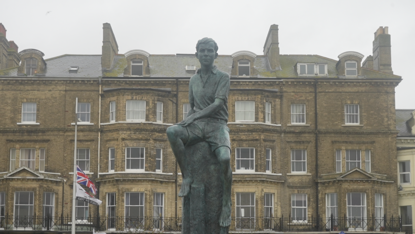 The statue of Benjamin Britten in front of a building divided into multiple properties. A flag pole with the Union Jack flag is also in the background.
