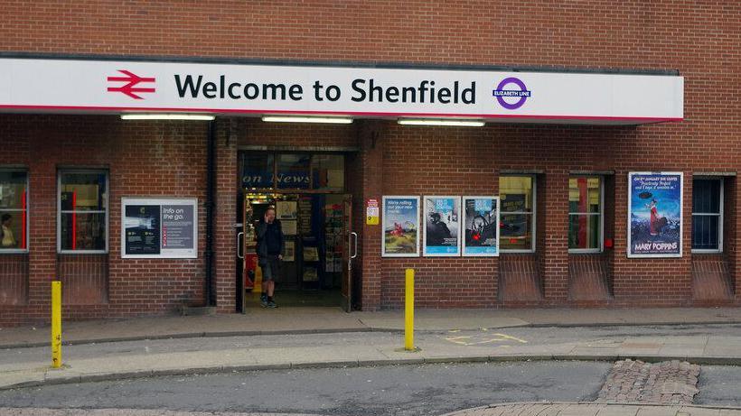 Exterior of Shenfield station. A man walks out of the entrance doors and is talking on his mobile phone. The building has advertising posters on its brick walls. 