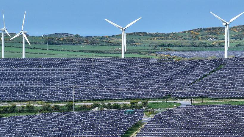 Several fields obscured by hundreds of solar panels with 4 wind turbines in the background and rolling hills further in the distance. 