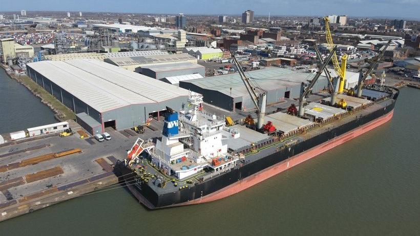 A large ship is moored next to the docks, with a number of warehouses shown and the city in the background.