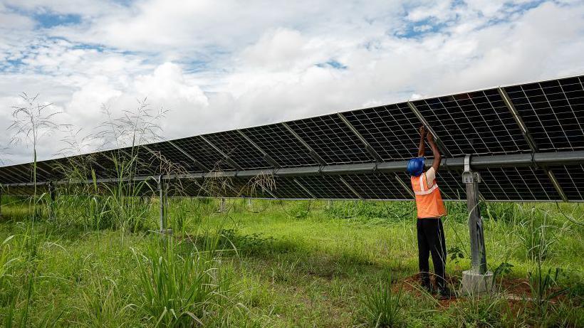 A man in hi vis and a hard hat reaches up to a large solar panel in a field.