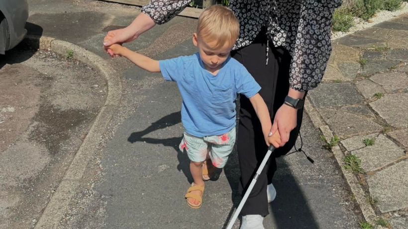 A young boy wearing shorts and a blue t-shirt, walking with a metal stick in his left hand. He is walking along a pavement helped by a woman behind him.