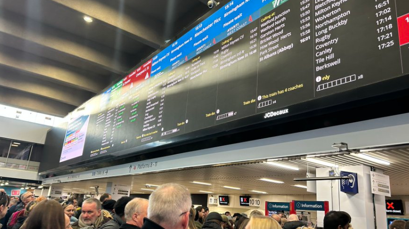 A large display in a station shows different rail services and their times. There are people stood under the display looking at the information.