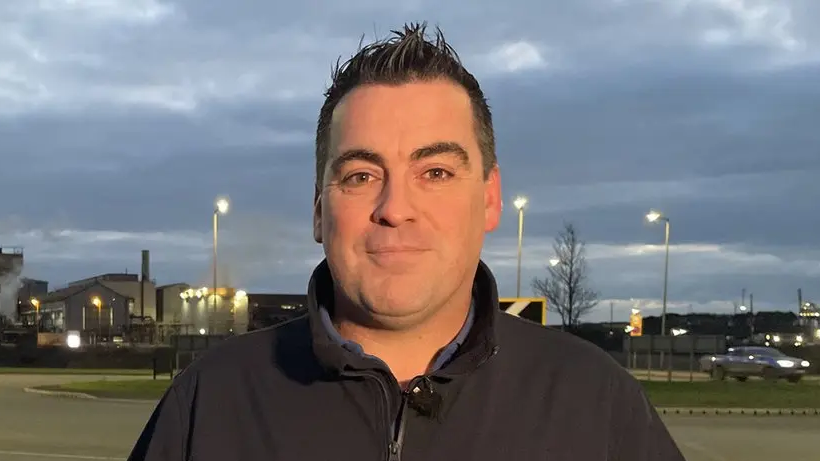 Shaun Spencer, a former worker at Tata's Port Talbot steelworks - which is visible behind him. He has short dark hair and is wearing a black fleece.