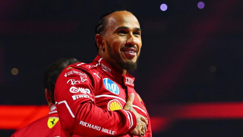 Lewis Hamilton holds his right hand across his chest while smiling at the F1 2025 season launch event. He is wearing the red Ferrari uniform 