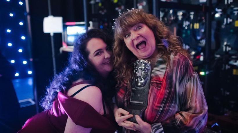 Janey Godley with daughter Ashley Storrie in November 2019. Ashley stands side on leaning into her mother's shoulder, smiling - she wears a red velvet dress and has long dark curly hair. Godley is dressed in plaid with long curly brown hair and is holding a Bafta award - she has her mouth open in celebration