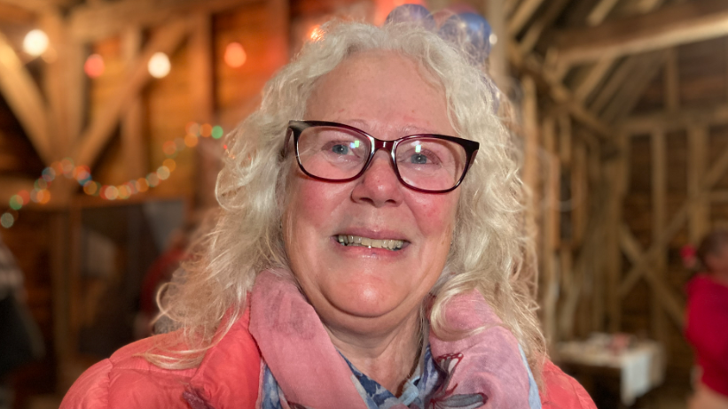 Dawn Briggs is in the middle of the image looking towards the camera smiling. She has long blonde curly hair, and is wearing brown glasses and a pink coat.