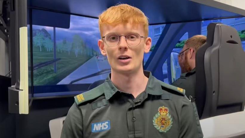 A young man with red hair and round wire-rimmed glasses in a green paramedic uniform, with NHS badge and West Midlands Ambulance Service badges. Behind him is a man sitting in a fake drivers seat in front of