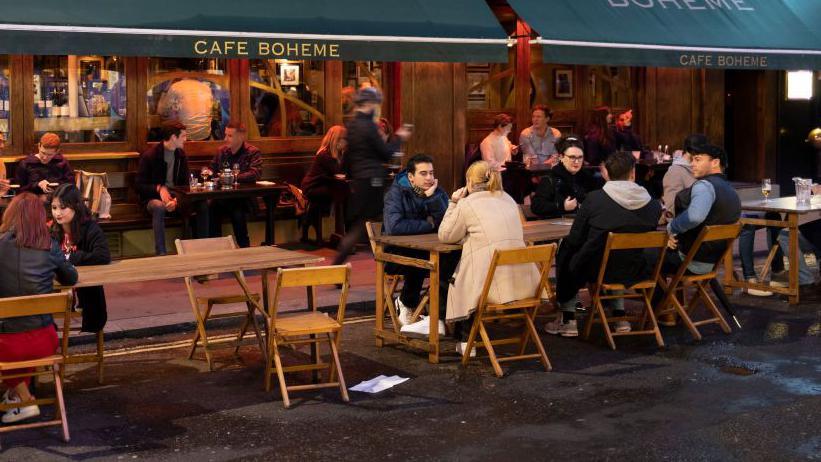 People seated at wooden tables outside Café Boheme under large green awnings at night, with a waiter serving drinks and pedestrians passing by.