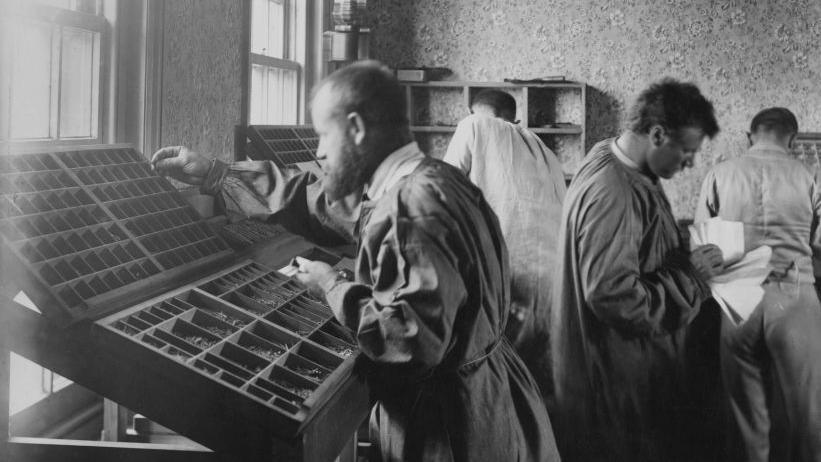 A black and white photo of Vladimir Tchertkoff working in the compositors' room of the Free Age Press in Southbourne. He is wearing a belted coat and standing in front of a wooden tray with dividers. Three other men are working in the background.