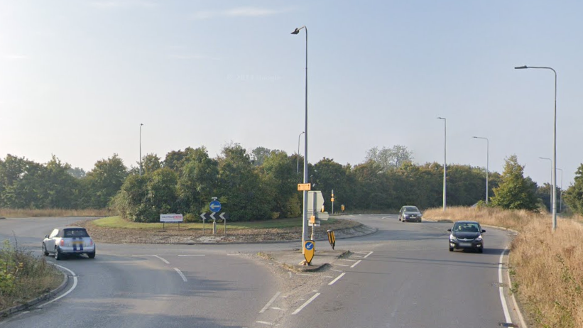 A Google streetview image of a roundabout with trees on it. There are three cars being driven around it. There are lampposts dotted around the roundabout and dry grass. It is sunny and the picture has clearly been taken in summertime.