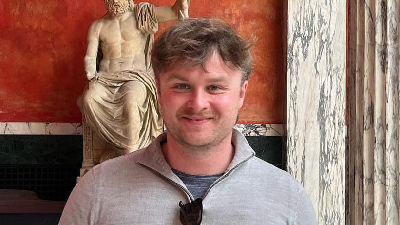 Hadley Sharman smiles at the camera. He has brown hair and a brown beard. He wears a three-quarter zip grey jumper with a dark grey top underneath. A Greek statue can be seen behind him.