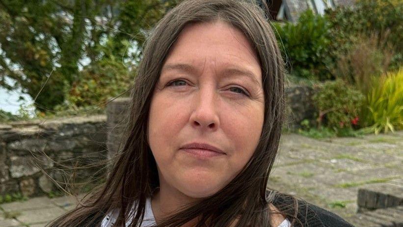 A woman with long dark brown hair is looking at the camera. She is wearing a white top with a black cardigan. She is standing on a paved area with trees and the sea blurred out behind her.