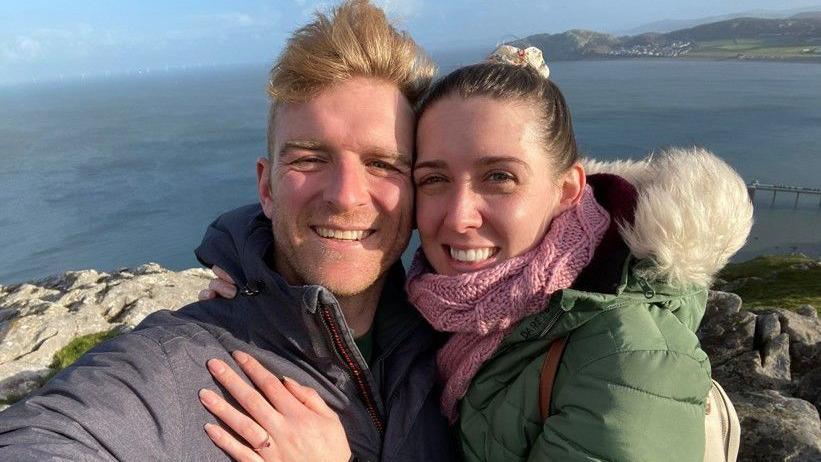 A selfie of Mick Thurston and Vicky Green embracing while out on a walk near the sea. Ms Green is wearing an engagement ring. Both are smiling.
