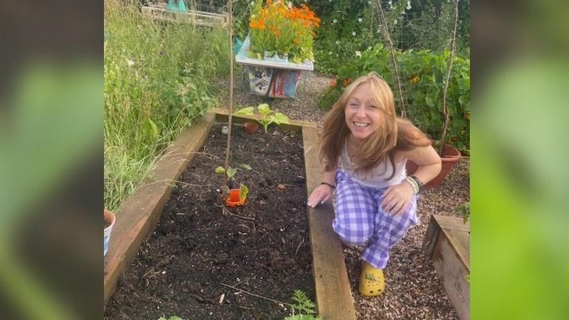 Laura is on one knee next to a rectangular piece of wood which is full of soil with a few plants in. She has blonde hair, a white top, and purple and white chequered trousers on. 