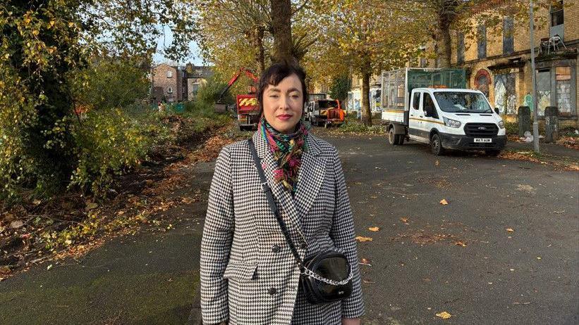 A woman with brown hair is standing in the street with trees and boarded up properties behind her. She is wearing a black and white dogtooth patterned coat with a black handbag over her shoulder.