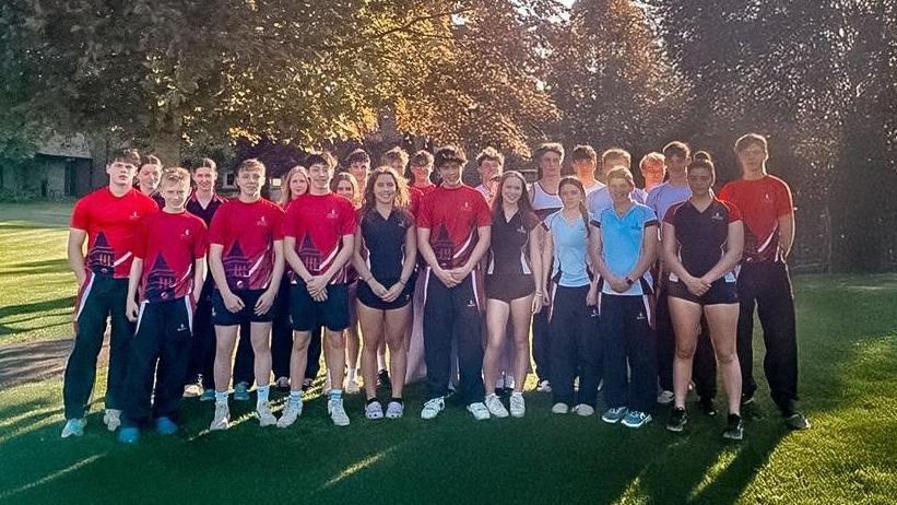 A group picture of students in sports clothes. It is sunny, they are standing under a tree.