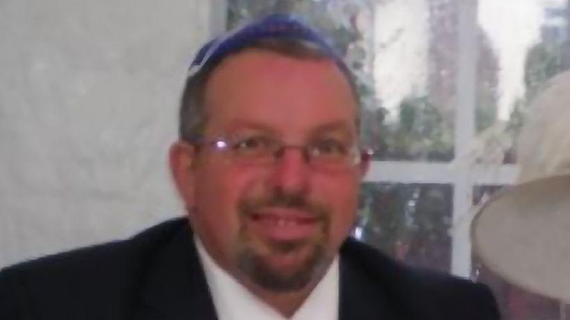 A man smiles at the camera. He is wearing a blue kippah on his head and glasses, and has a goatee. He is wearing a black suit jacket and a white shirt. Behind him is a blurred window. 