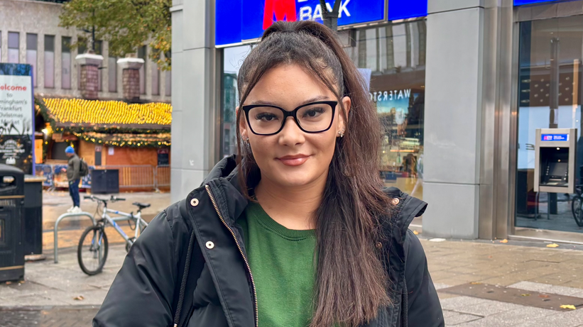 A woman with black hair, glasses, a green jumper and a coat looking into the camera.