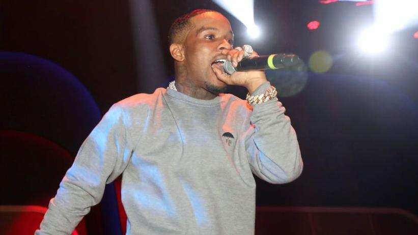 Tory Lanez performs on stage. He is wearing a grey jumper and is singing into a mic. He stands in front of black background. 