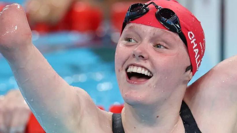 Ellie Challis. She is in the pool wearing a red swimming cap and black swimming googles on her head. Challis is in a swimming pool and smiling with her arms in the air