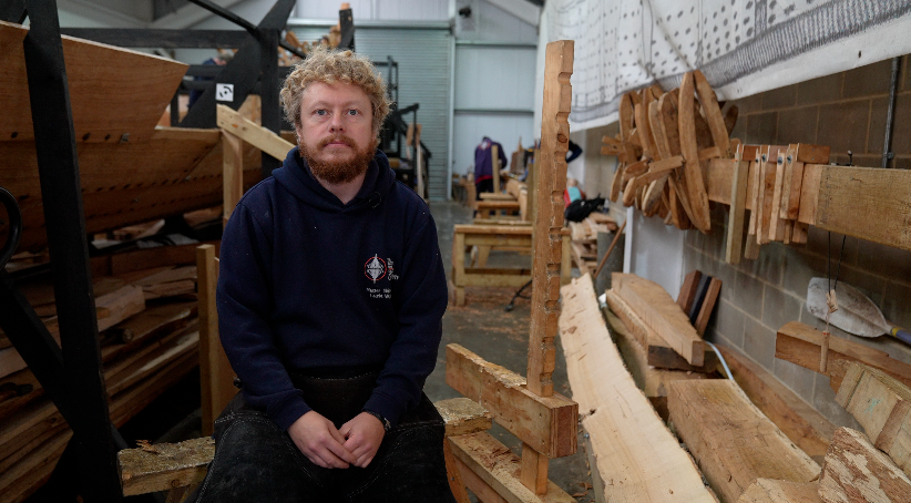 Laurie Walker smiles at the camera as he sits within the ship's workshop. He has blonde curly hair and a red beard. Part of the ship can be seen behind him as well as some wood planks. He wears a blue hoodie. 
