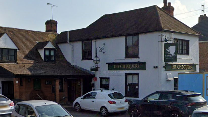 Street view of the pub with a number of cars parked next to it. It is a white building with a green sign with 'Chequers' written on it in gold embossed lettering.