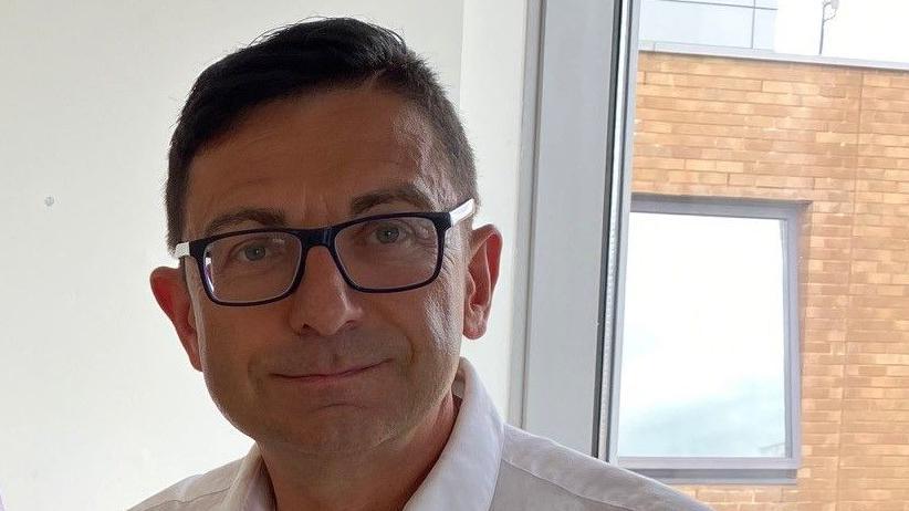 A smiling Dr Vaclav Dosta looks directly at the camera as he is photographed wearing a white shirt, and black-rimmed spectacles. He has short dark hair. In the background is a window, which shows brickwork outside the hospital and another window.