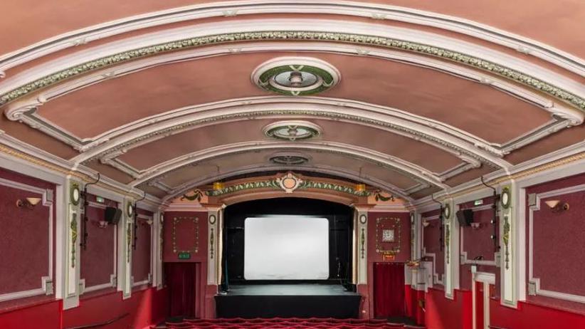 The inside of a old fashioned cinema hall. The ceilings are low and the walls are white, pink and red 