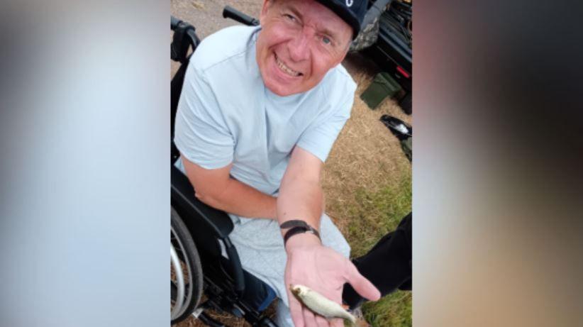 In a close-up photograph, Richard Rudd is seen leaning towards the camera and in his palm he has the fish he has just caught at the lake. He is wearing a grey T-shirt and tracksuit bottoms and a dark blue baseball cap with New York written on it in white.
