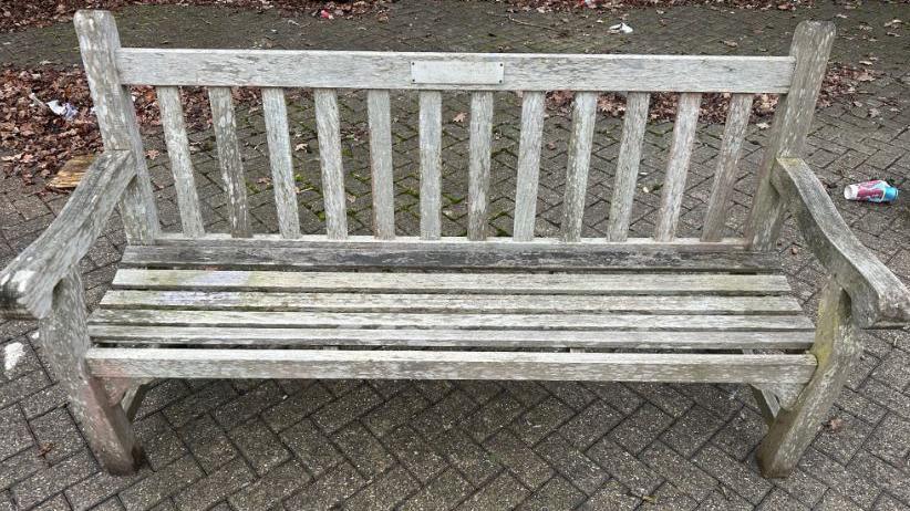 A brown wooden bench that has been bleached by the sun and the elements. A small plaque is top centre of the backrest, the words cannot be made out. The bench sits on grey pavement tiles