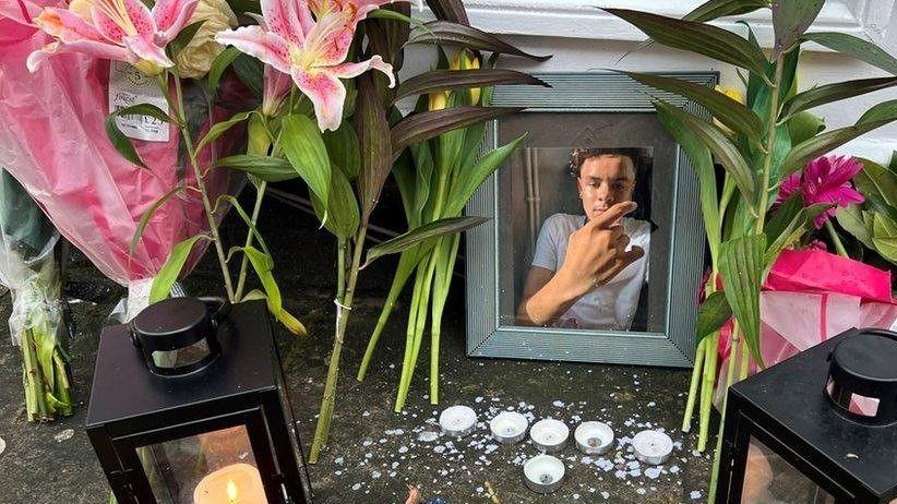 A shrine set up to Darrian near the scene of where he died. There are two black lanterns with candles inside, some melted tealights on the floor, a framed picture of Darrian and lots of bouquets of flowers.