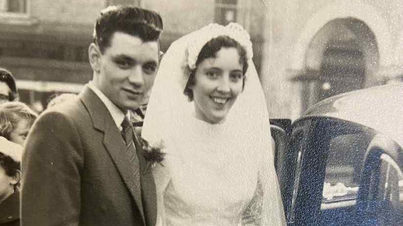 A black and white photo shows a wedding day. A man and a woman stand close together: the man in a formal suit, the woman in a traditional white wedding gown with a veil. They are next to a car, and there are people and buildings visible in the background.