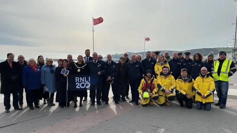 People gather for the RNLI 200 ceremony on Douglas Promenade. Some people wear yellow RNLI coats, an flag is held up by the city's Mayor. It is a hazy day.