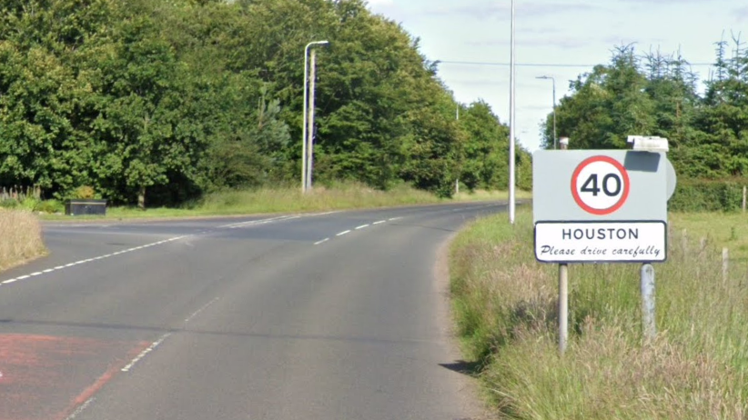 A country road with grassy fields on either side with trees in the distance. A sign says the speed limit is changing to 40mph and it also reads "Houston, please drive safely"
