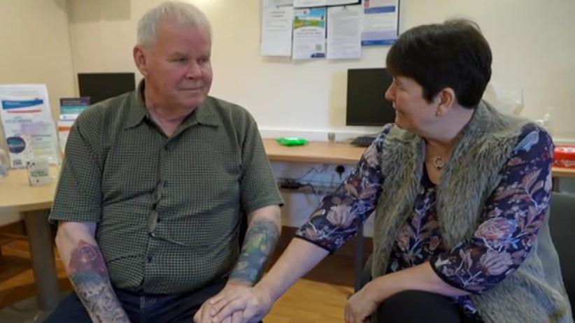 Two people sat side by side in a room with a wooden floor and beige walls. Shane Game is sat on the right of the image; she is wearing a purple top with a floral pattern and a grey fluffy waistcoat. She is looking at Mark Scott, on the left and wearing a green button-up short-sleeved shirt. Shane is holding Mark's hand and he is looking at her while she speaks. 