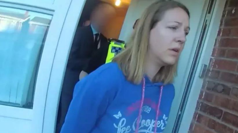 Lucy Letby wearing a blue hoodie leaving a house in handcuffs after her arrest with a police officer behind her in the front doorway